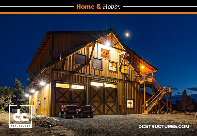 Barn-style home illuminated at night with warm lights features a wooden exterior and garage with living quarters. Large front doors and a side staircase add charm. A starry sky and bright moon serve as the backdrop, while two ATVs sit parked in front. Website link in the lower right corner.