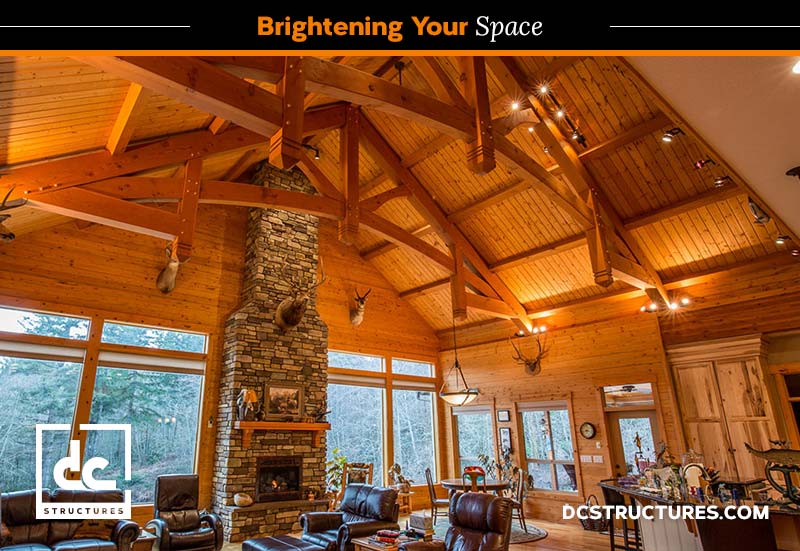 A spacious wooden interior features a high, vaulted ceiling with exposed beams. A large stone fireplace is central, set against a grand window wall. Decor includes mounted deer heads and rustic furnishings. Text reads "Brightening Your Space" and "DCStructures.com".