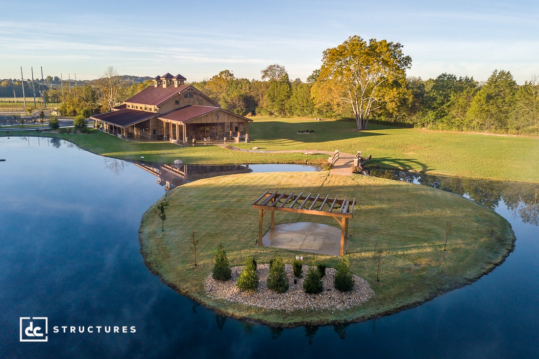 Aerial view of a rustic wooden building next to a large pond featuring an outdoor wedding pavilion on a small round island with a pergola. The surrounding landscape includes lush grass, trees, and a clear blue sky. Logo in the corner says DC Structures.
