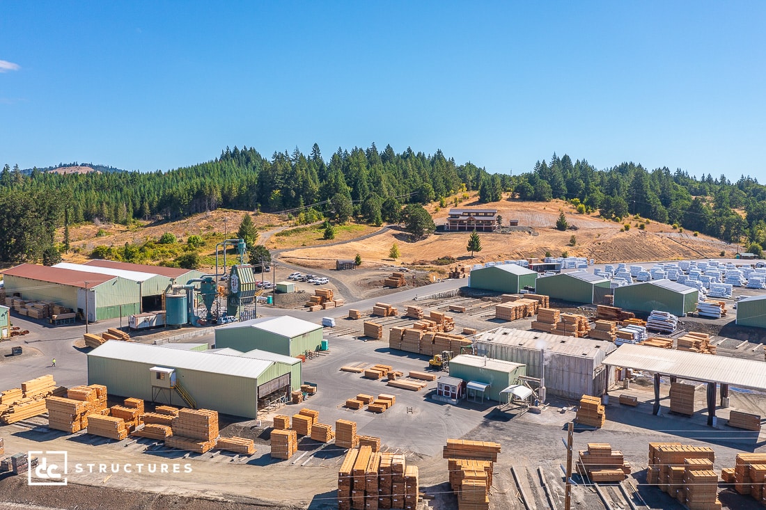 Aerial view of a lumber yard nestled in the historic old mill district, with stacks of wood and several warehouses. Surrounding the yard are trees and hills under a clear blue sky.