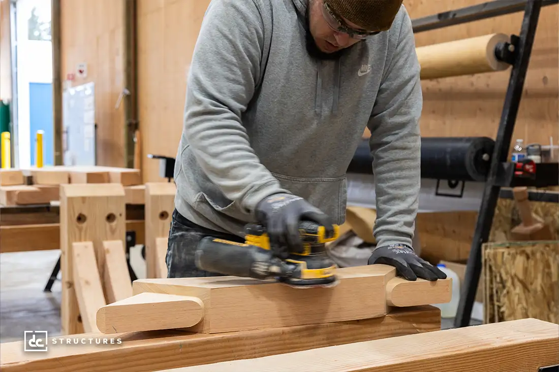 A person wearing a gray hoodie and gloves is using a power tool to sand a piece of wood inside a workshop, surrounded by wooden beams and cylindrical logs. The scene resembles the meticulous crafting found in post-and-beam kits.