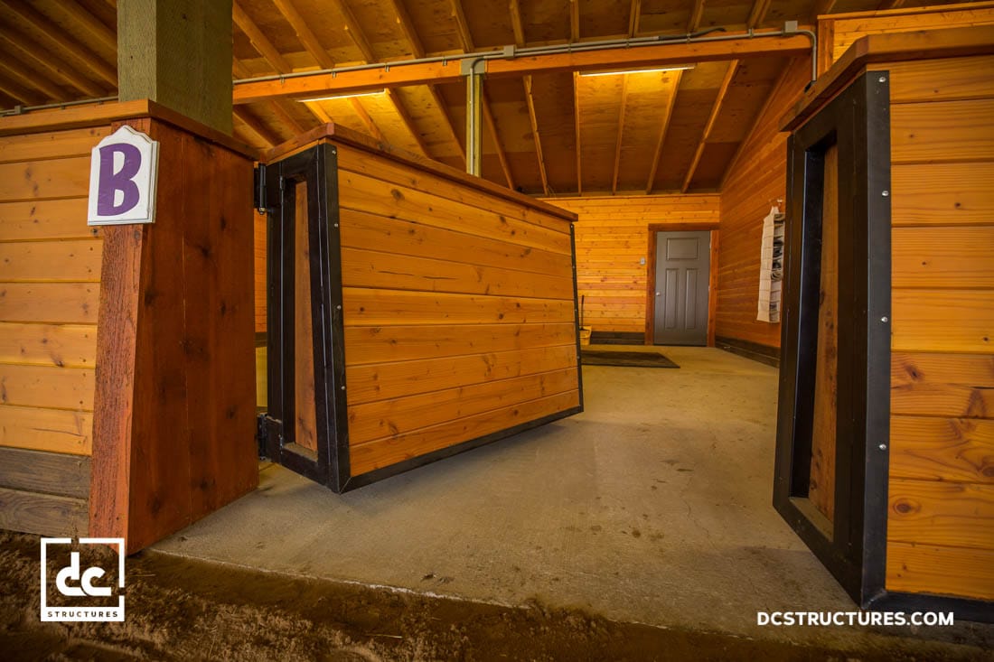 The interior of a wooden horse stable with a partially open gate showcases natural wood walls and ceiling. A sign with the letter "B" is mounted on a post, while the concrete floors offer durability. Perfectly designed for horse arena kits, the DC Structures logo adorns the corners seamlessly.