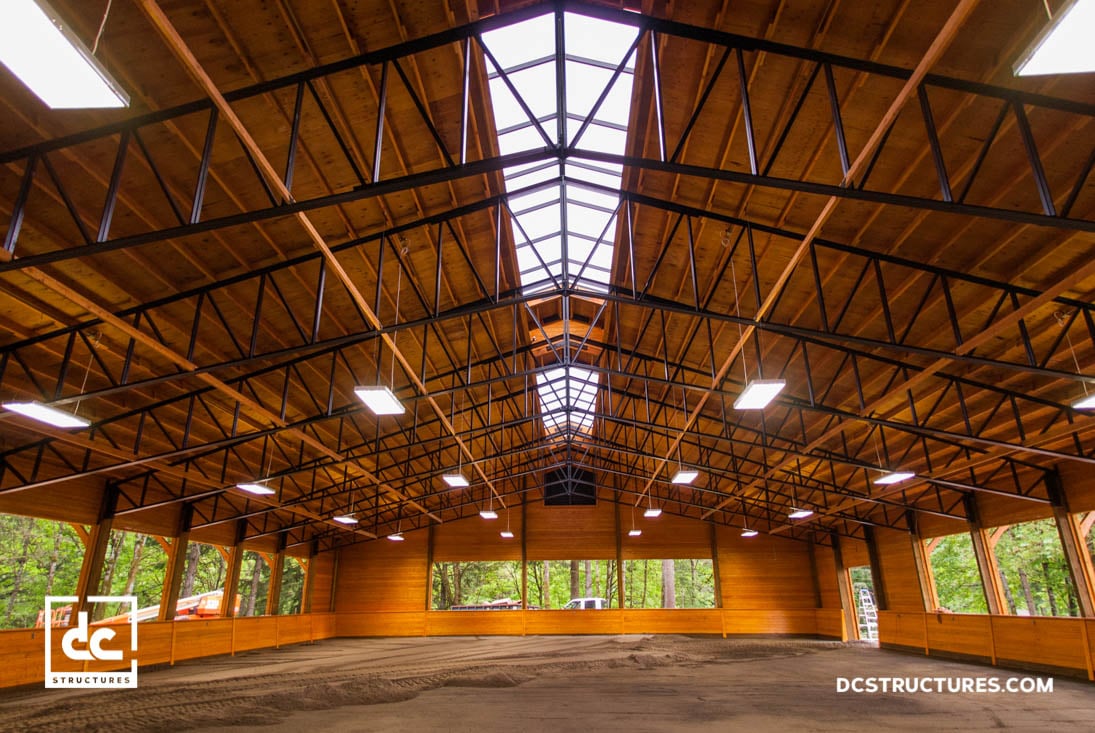 The interior view of a large wooden building, possibly crafted using horse arena kits, features a vaulted ceiling with exposed beams. Sunlight pours in through a skylight, complemented by artificial lights overhead. With its dirt floor and trees visible through the windows, it exudes rustic charm.