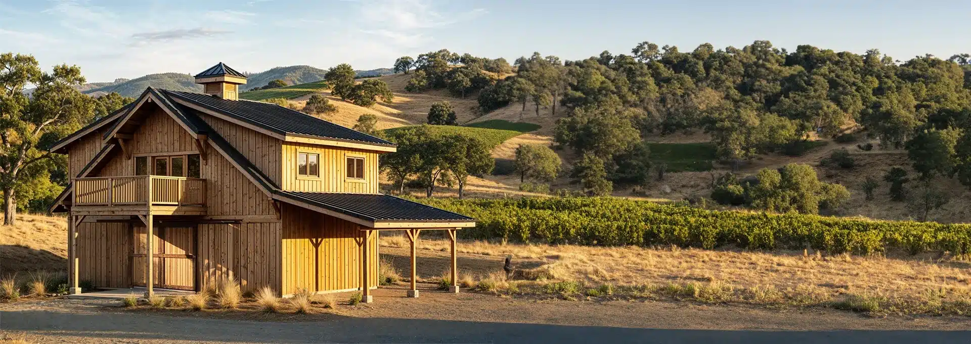 A wooden two-story barn with a balcony sits in a sunlit rural landscape, surrounded by dry grass, hills, trees, and a vineyard.
