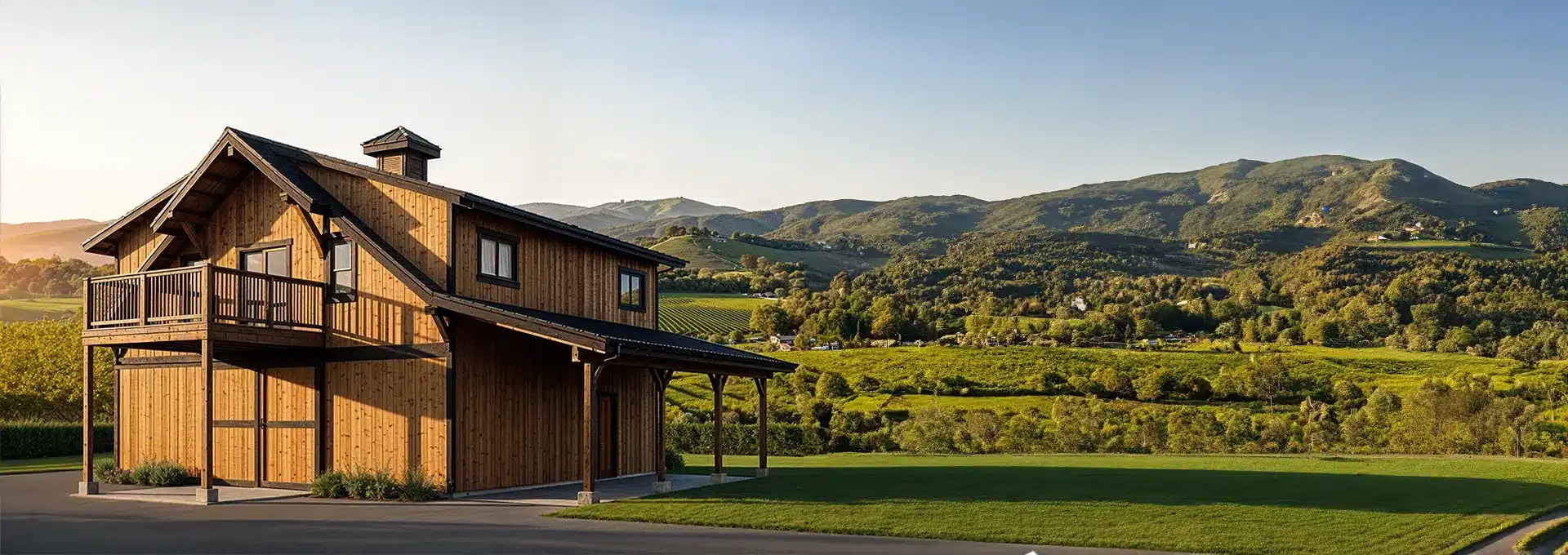 A wooden house with a balcony stands on a paved area, overlooking green hills and distant mountains under a sunset sky.