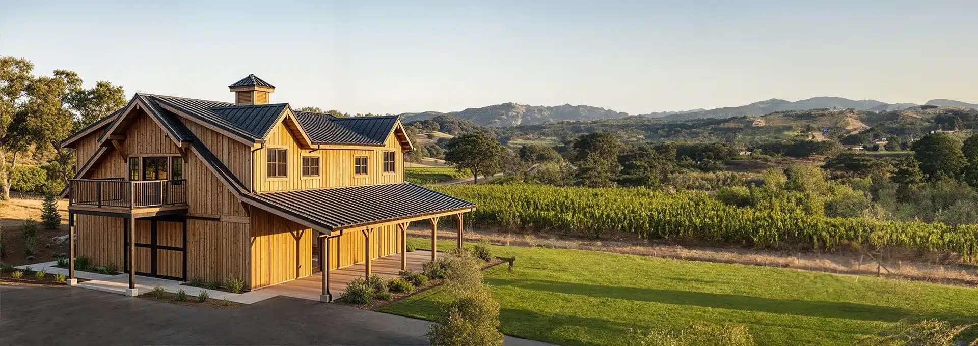 A large wooden barn with a metal roof stands beside green fields and vineyards, with hills and mountains in the background.