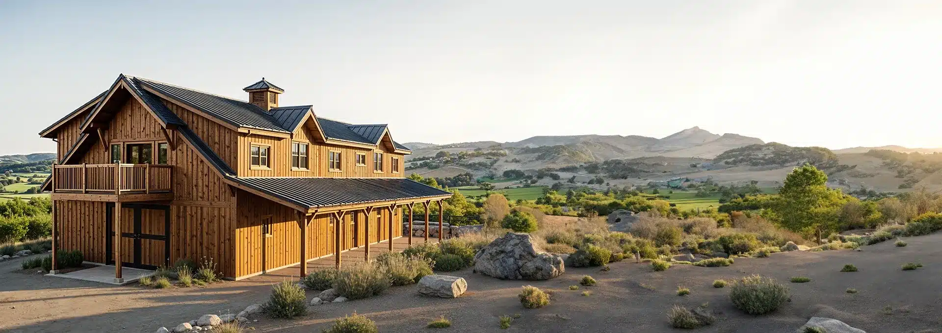 A rustic wooden barn with a balcony amid a dry, shrub-dotted landscape, rolling green fields, and mountains under a sunny sky.