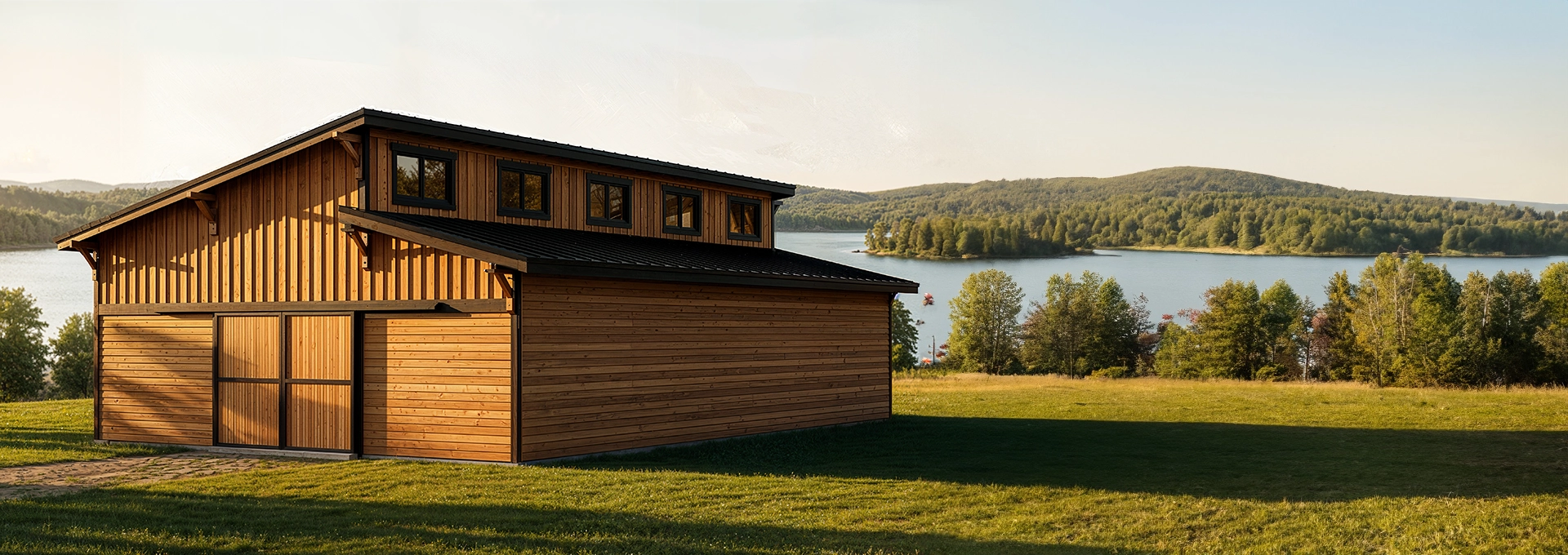A modern wooden barn with large windows stands on a grassy field overlooking a scenic lake and forested hills under a clear sky.