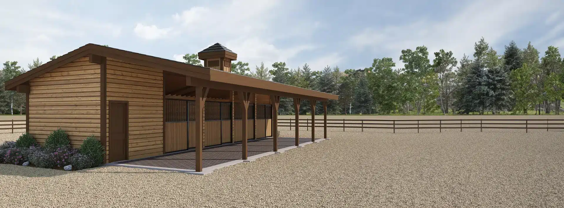 A wooden horse barn with a covered porch stands on gravel, surrounded by a fenced area and trees under a partly cloudy sky.
