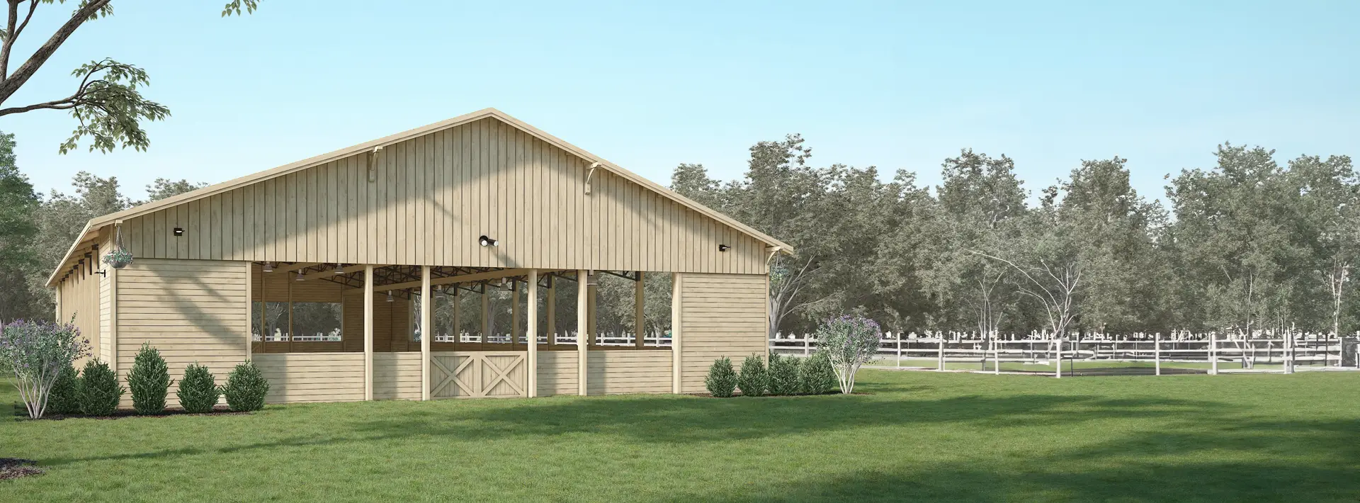 A large wooden barn with open sides stands on a grassy lawn, surrounded by greenery and trees, with a white fence and blue sky.