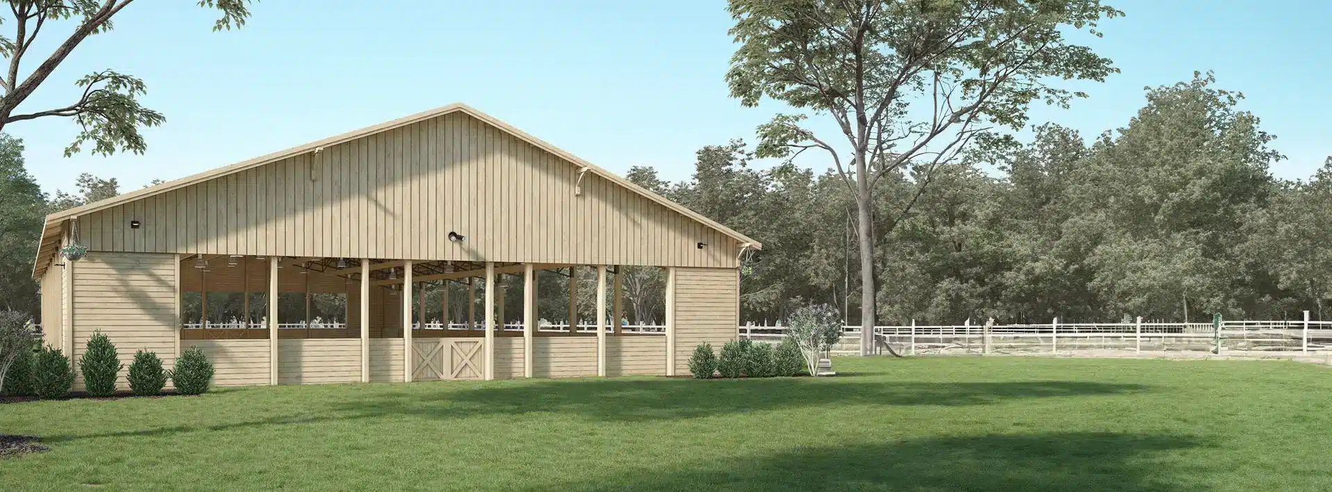 A light wooden barn with open sides stands on a green lawn, surrounded by trees and bushes under a clear blue sky. A white fence runs through the background near the edge of a wooded area.