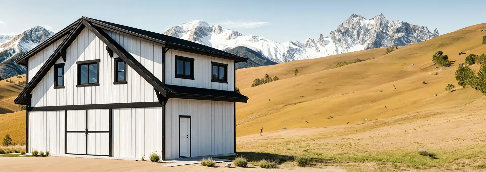 A modern white barn-style house with black trim stands in a golden field, with snow-capped mountains and a clear blue sky behind.