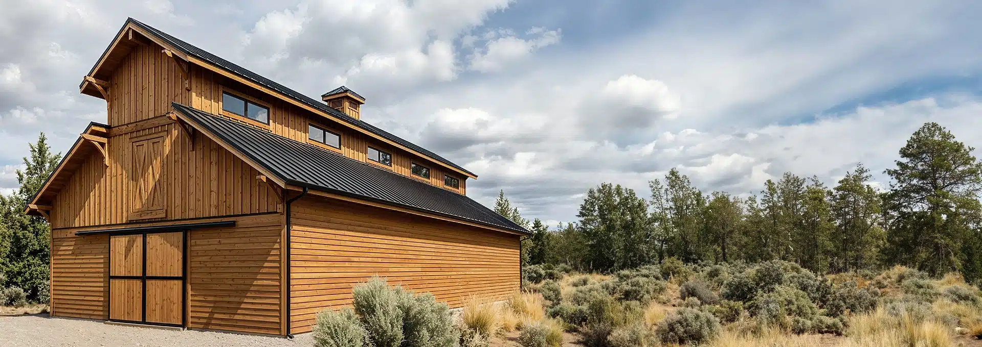 A large wooden barn with a black metal roof stands near shrubs and trees under a partly cloudy sky in a rural landscape.