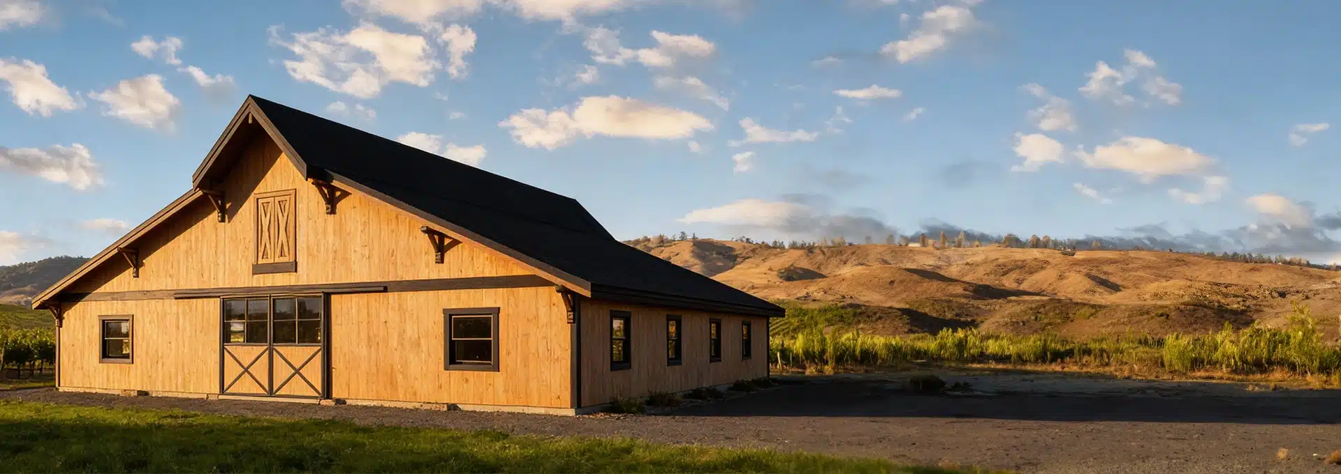 A large wooden barn with a black roof stands on gravel, surrounded by green fields and dry hills under a blue sky with clouds.