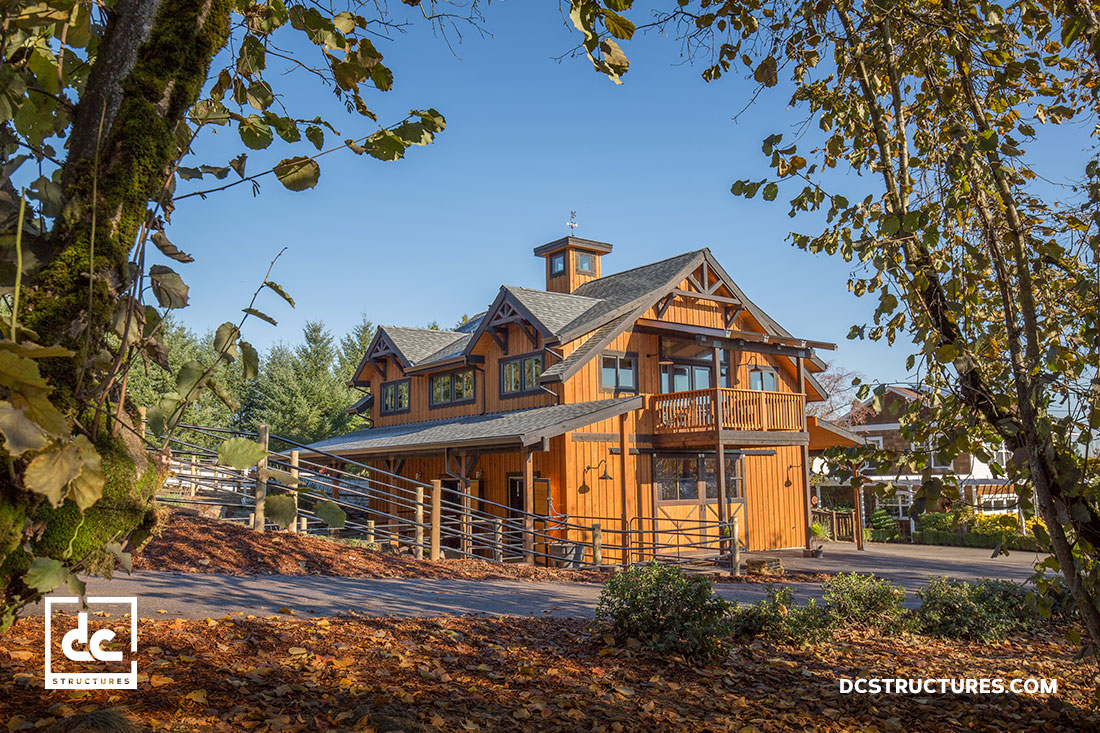 A two-story wooden barn surrounded by trees and autumn leaves under a clear blue sky. The structure features a balcony and rustic design. The image includes the logo and website of DC Structures in the corners.