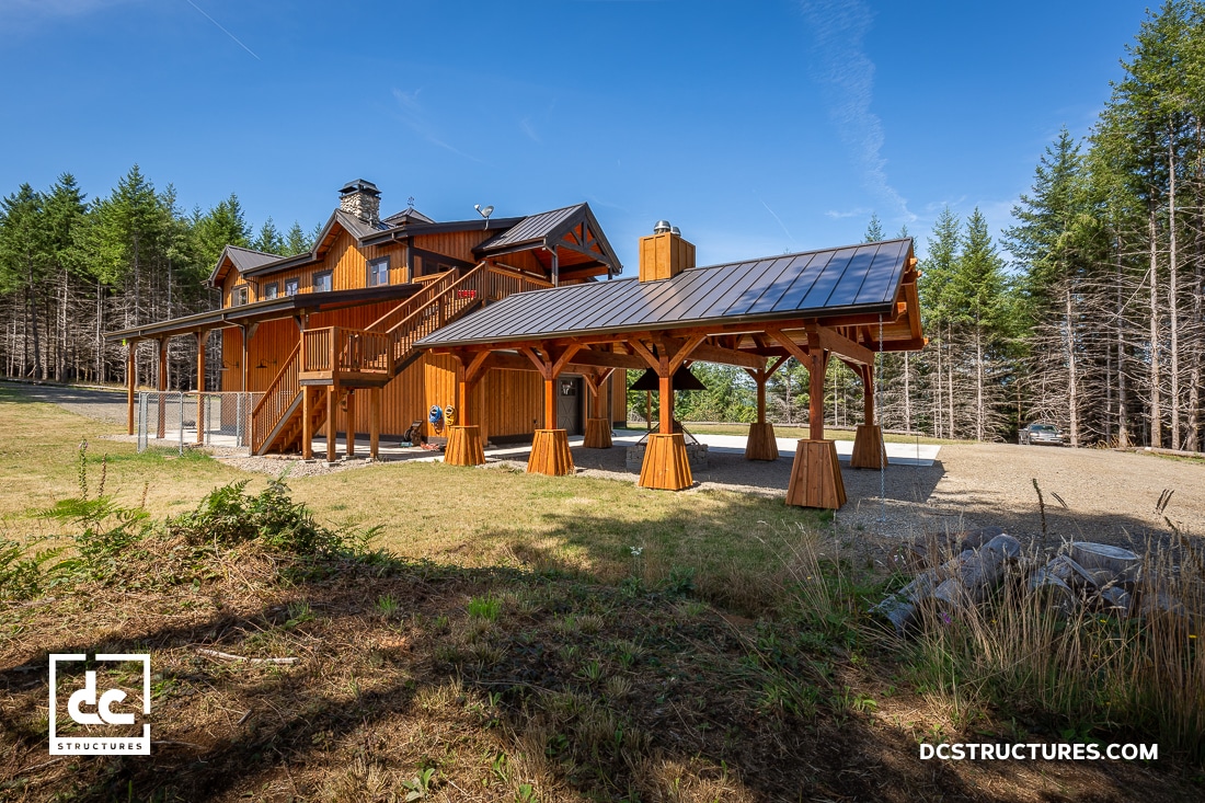 A large wooden house with a covered outdoor area stands on a grassy plot surrounded by pine trees under a clear blue sky.