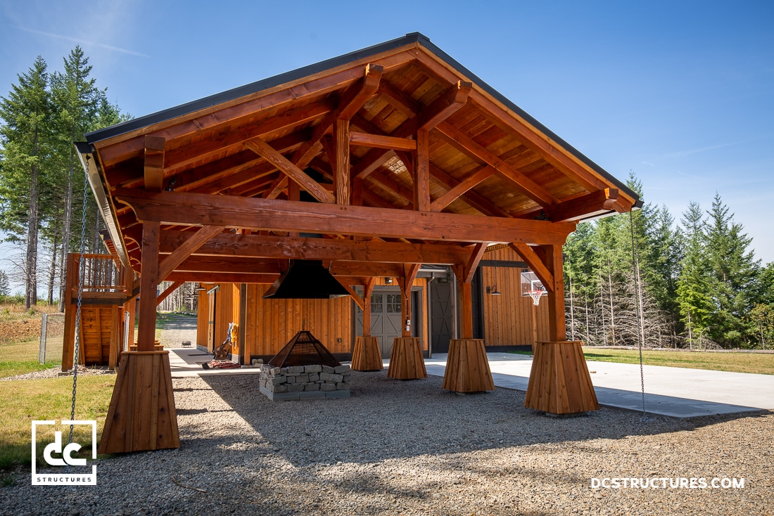 A wooden pavilion with exposed beams and a pitched roof stands over a stone fire pit on gravel near a wooden building and trees.