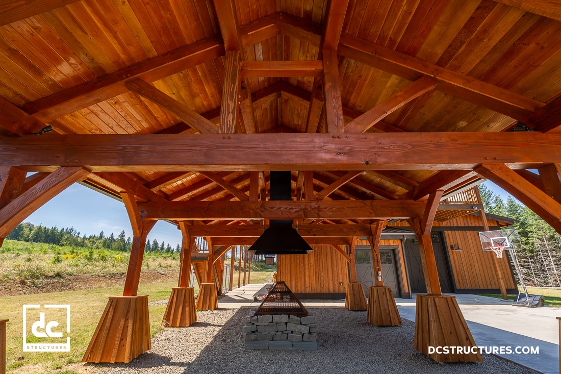 Covered outdoor wooden pavilion with exposed beams, a metal fireplace, picnic tables, and gravel floor. Greenery surrounds it.