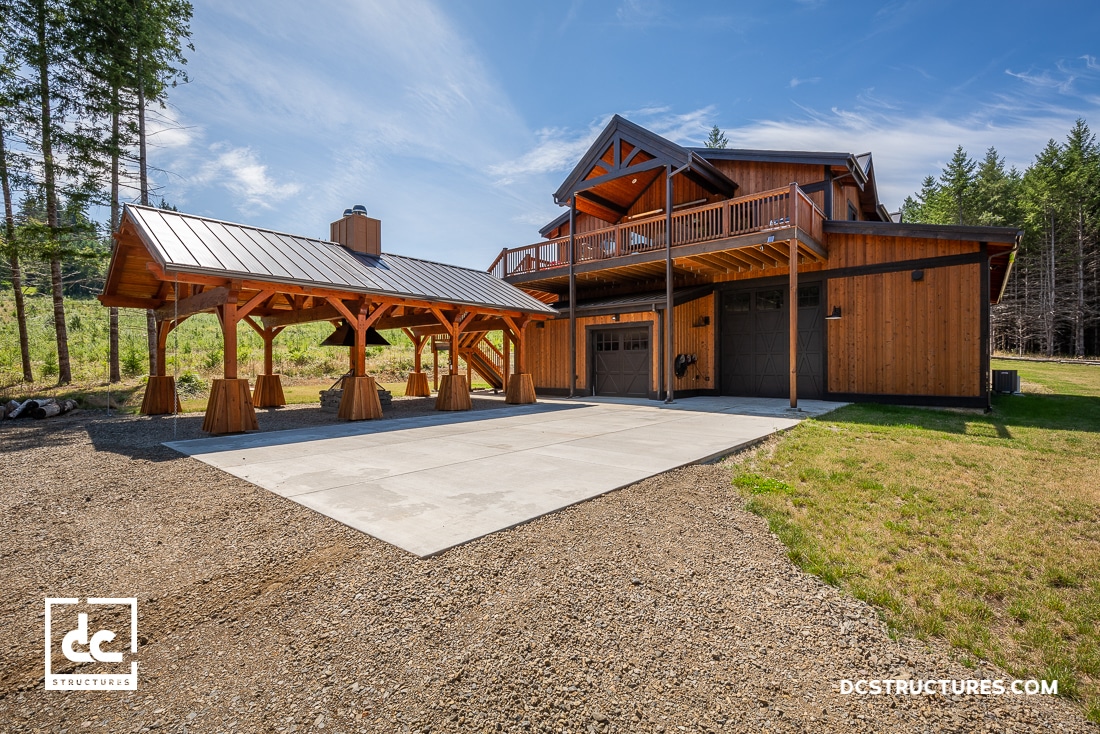 A large wooden barn-style home with an attached covered pavilion, a concrete driveway, and surrounded by grass, trees, and blue sky.