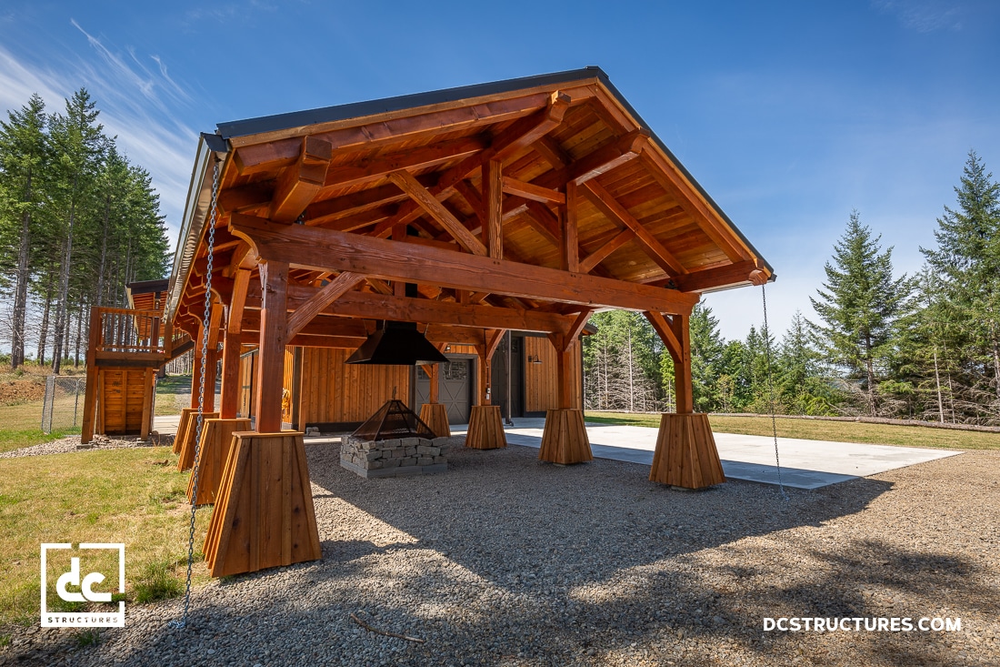 A wooden pavilion with exposed beams and a pitched roof sits on gravel next to a forest. A fire pit is located in the center.