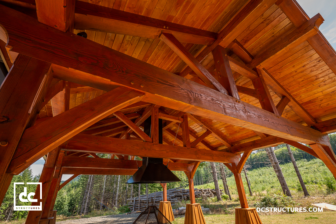 A close-up view of a wooden pavilion’s sturdy timber frame with a hanging fireplace, set outdoors among trees under a clear blue sky.