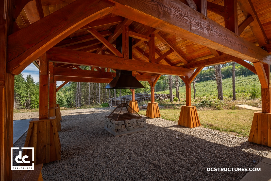 A wooden pavilion with a pitched roof shelters a central stone fire pit and metal chimney, surrounded by gravel, trees, and open sky.