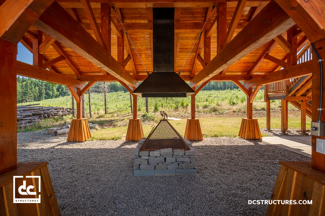A wooden outdoor pavilion with a central grill, chimney, stone base, and gravel floor, surrounded by green fields and trees. Sunlight highlights timber beams.