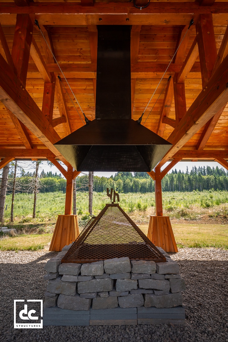 A covered outdoor fire pit made of stacked stones with a metal grate and large black chimney hood, under a wooden pavilion in a forested setting.