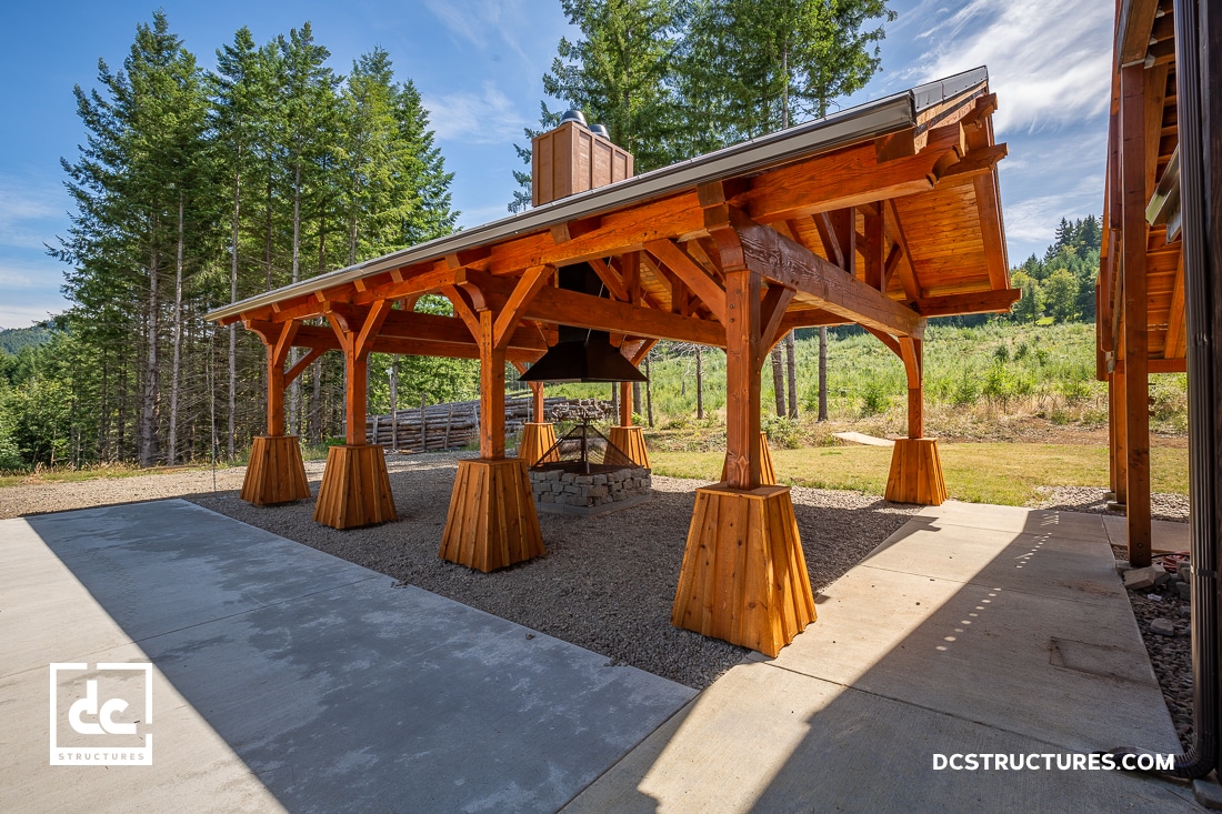 A wooden pavilion with a gabled roof, reminiscent of high-quality pavilion kits, stands on a concrete and gravel base in a forested area. Large timber posts rest on angled concrete footings, while tall trees and green hills provide a stunning backdrop.