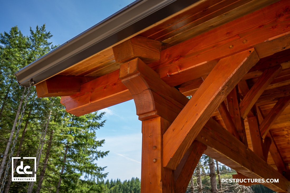 Close-up of a timber frame structure with sturdy wooden beams and joints under a roof, set against green trees and blue sky.