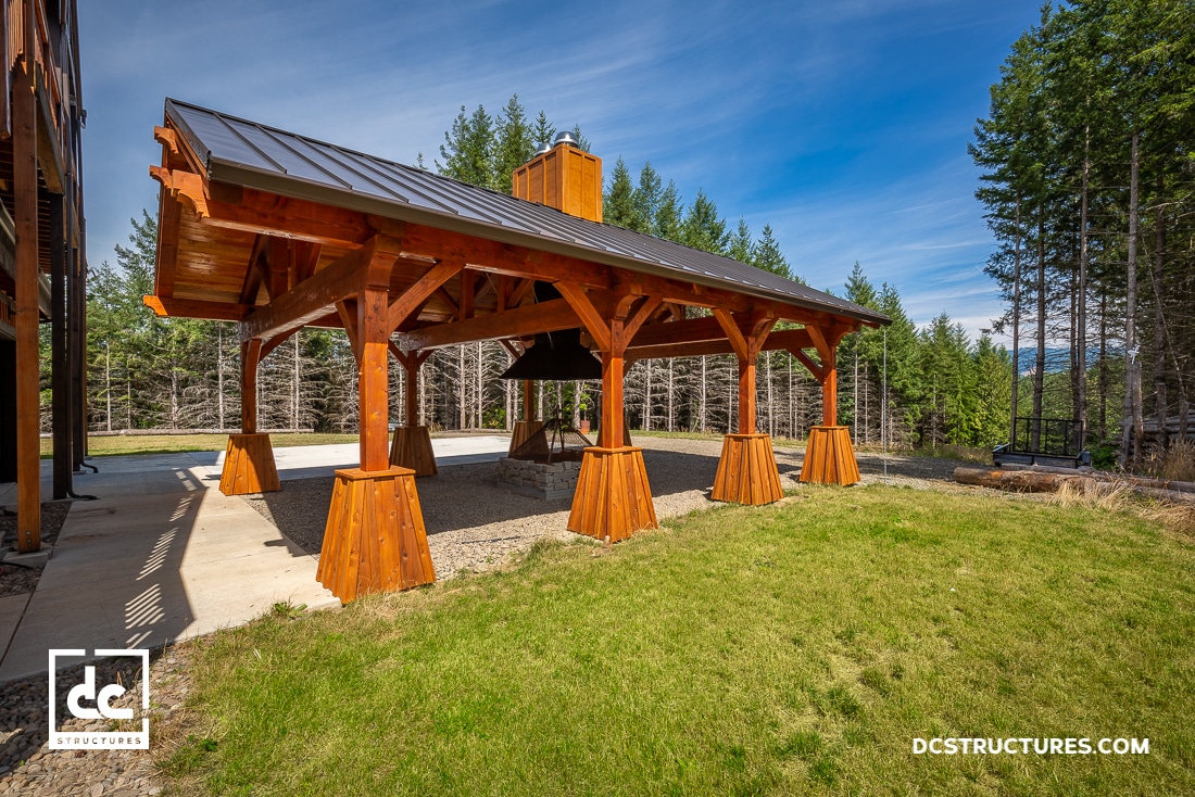 A wooden pavilion with a metal roof stands on a concrete and gravel base in a grassy area, surrounded by tall green trees under a clear blue sky.