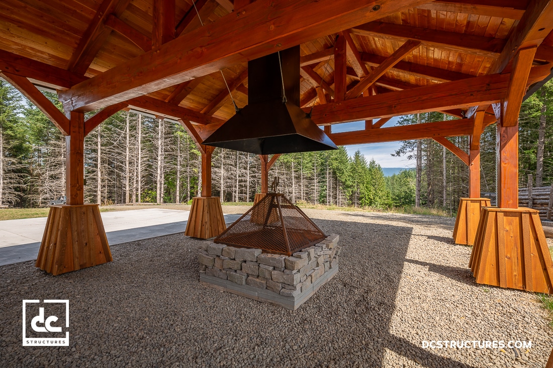 A wooden pavilion with an open beam roof shelters a stone fire pit with a large metal hood. Gravel, trees, and mountains in background.