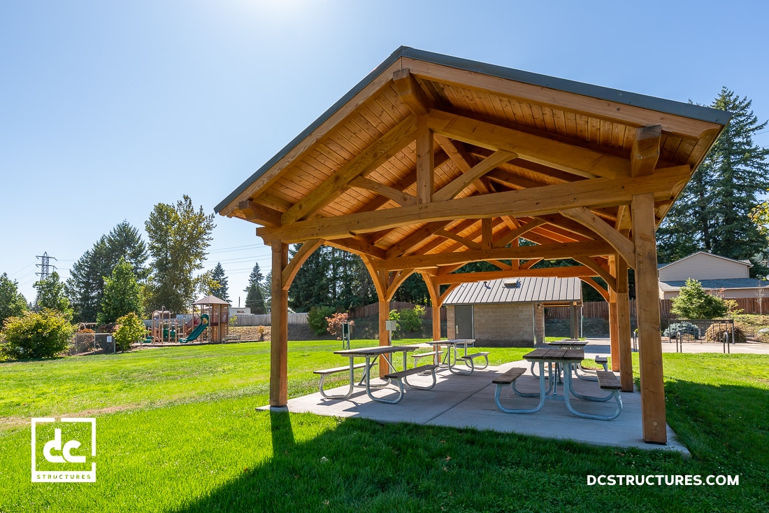 A wooden picnic shelter with a gabled roof stands on a grassy park lawn. A playground and building are in the background.