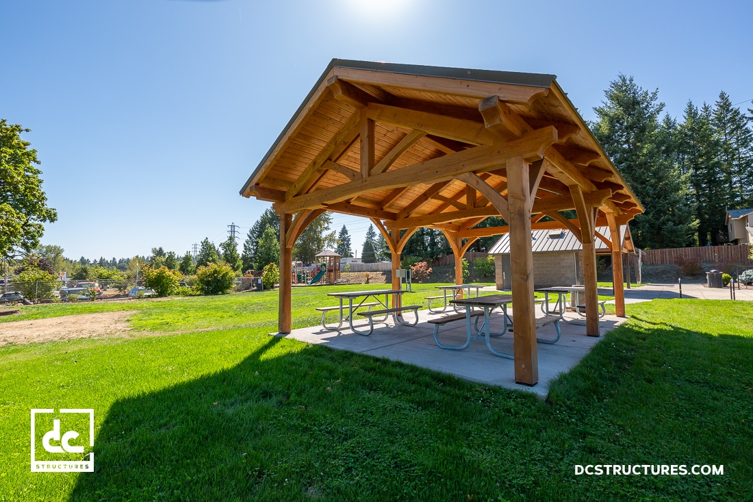 A wooden picnic shelter with a gabled roof stands on a concrete pad in a grassy park, with picnic tables underneath. Trees and a playground are in the background under a clear blue sky.