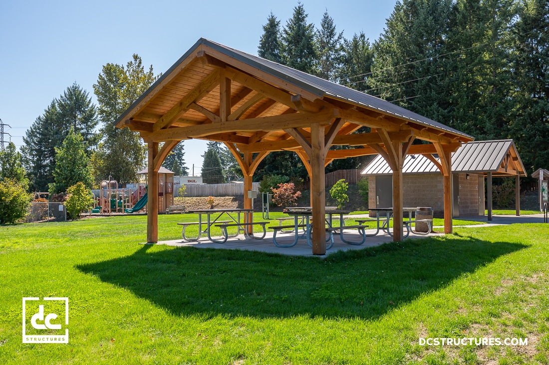 Wooden picnic shelter with a gabled roof in a park setting. The shelter covers a picnic table on a concrete slab. Playground equipment is visible in the background, surrounded by green grass and tall trees under a clear blue sky.