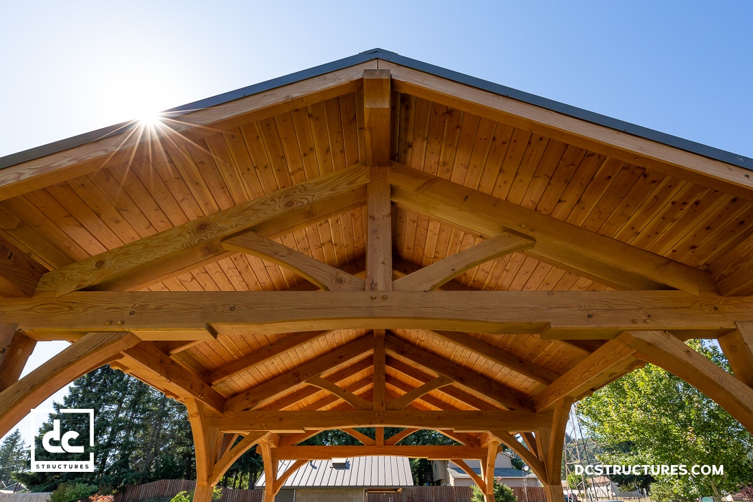 A wooden pavilion roof with exposed beams and trusses, photographed from below on a sunny day. Sunlight shines near the edge.