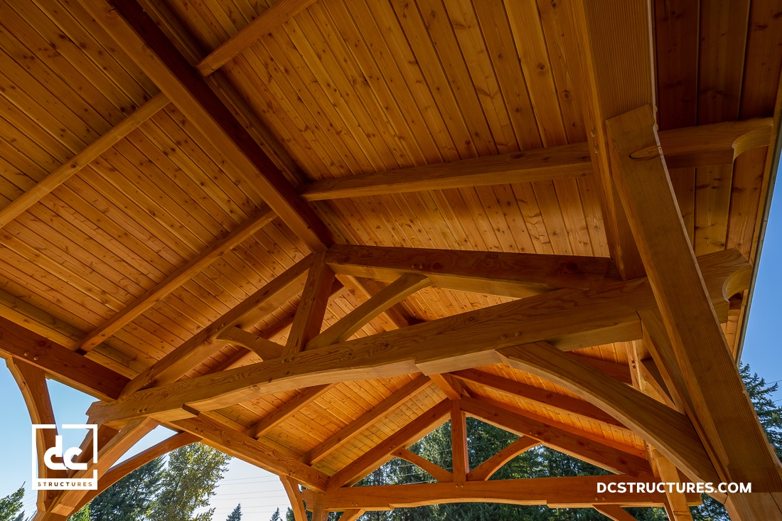 View of a timber frame pavilion roof with exposed wooden beams and tongue-and-groove ceiling, photographed from below with blue sky and trees at the edges.