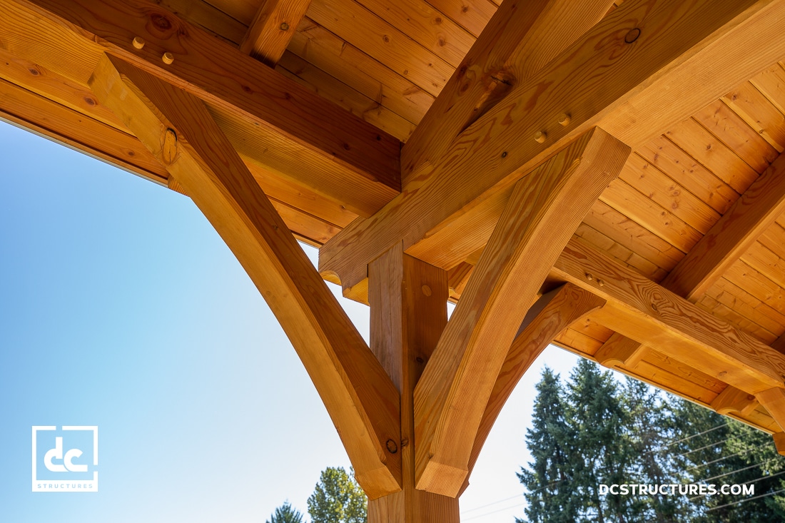 Close-up view of timber frame construction with large wooden beams and joinery, under a roof with blue sky and trees in the background.