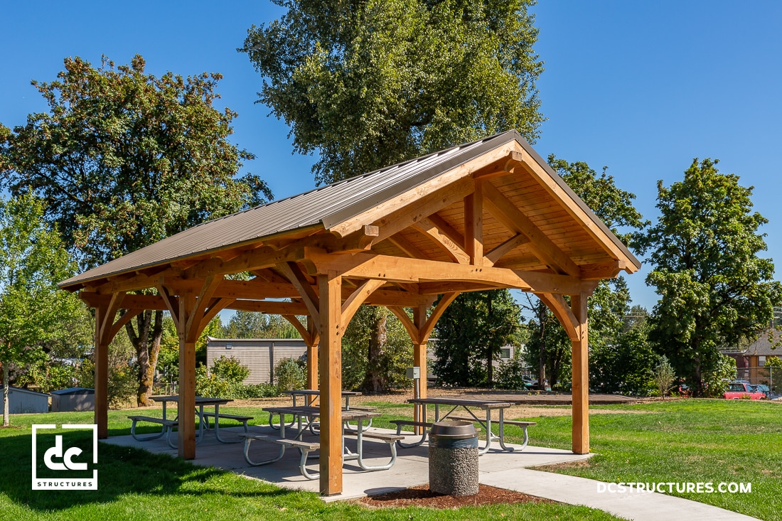 A wooden picnic shelter with a peaked roof stands in a grassy park, shading several picnic tables. Trees and blue sky are behind.