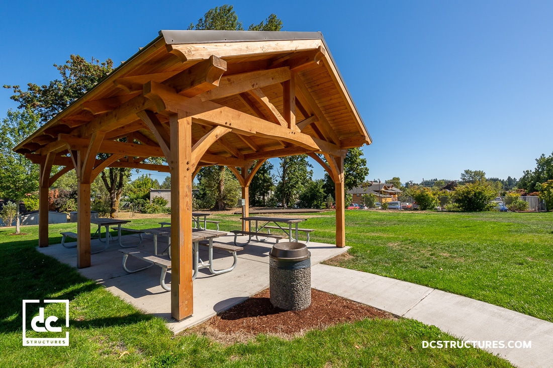 A wooden pavilion with picnic tables sits on a concrete pad in a sunny, green park. Trees and a playground are in the background.