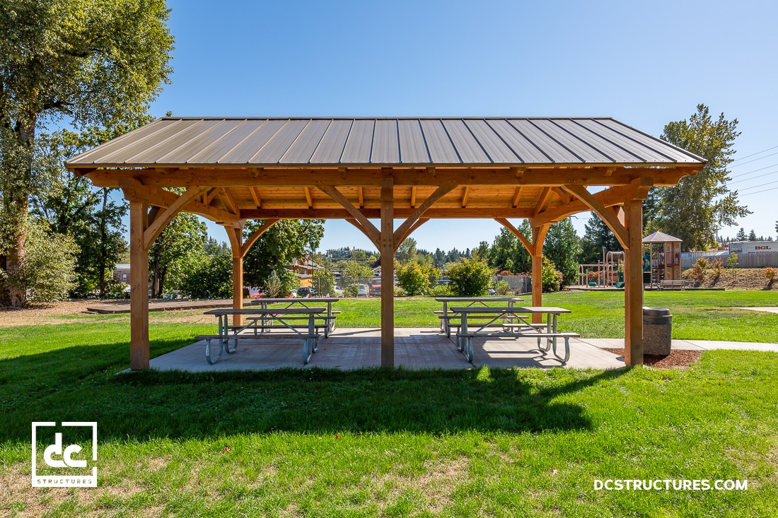 A wooden picnic shelter with a metal roof stands on a concrete pad in a sunny park. Two picnic tables are under the shelter, surrounded by green grass and trees. The sky is clear and blue.