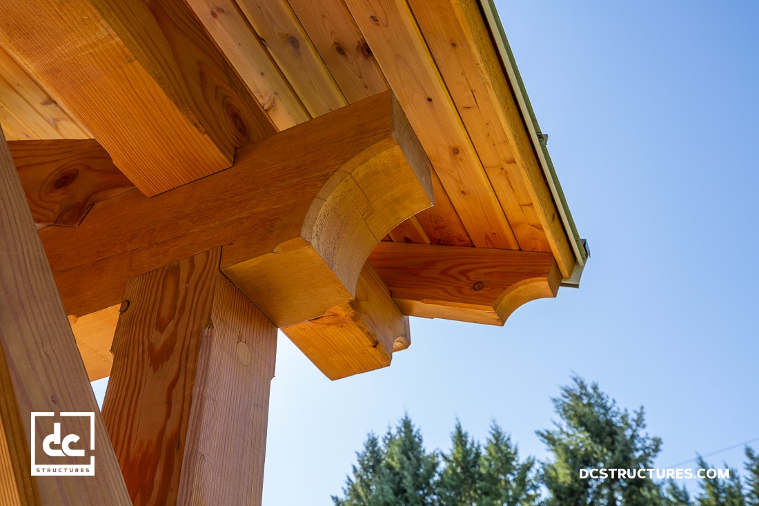 Close-up of a wooden pergola or pavilion roof corner, showing detailed joinery and beams against a blue sky with trees behind.