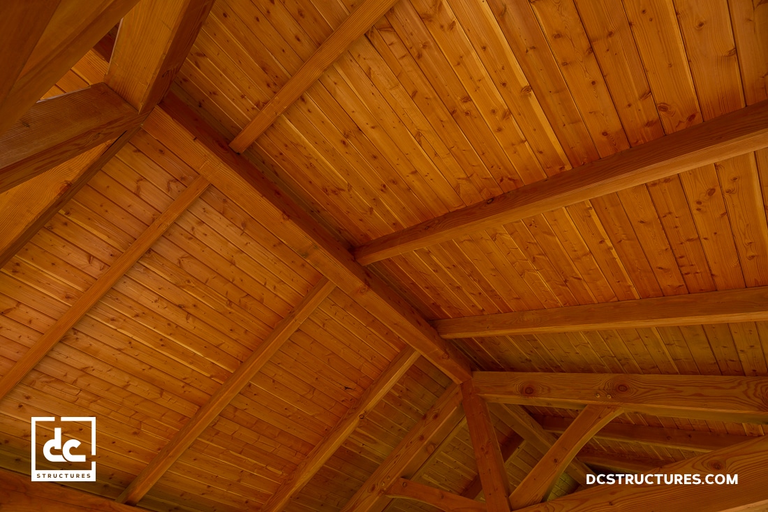 Angled view of a wooden ceiling with exposed beams and tongue-and-groove paneling. The warm, honey-colored wood shows detailed craftsmanship.