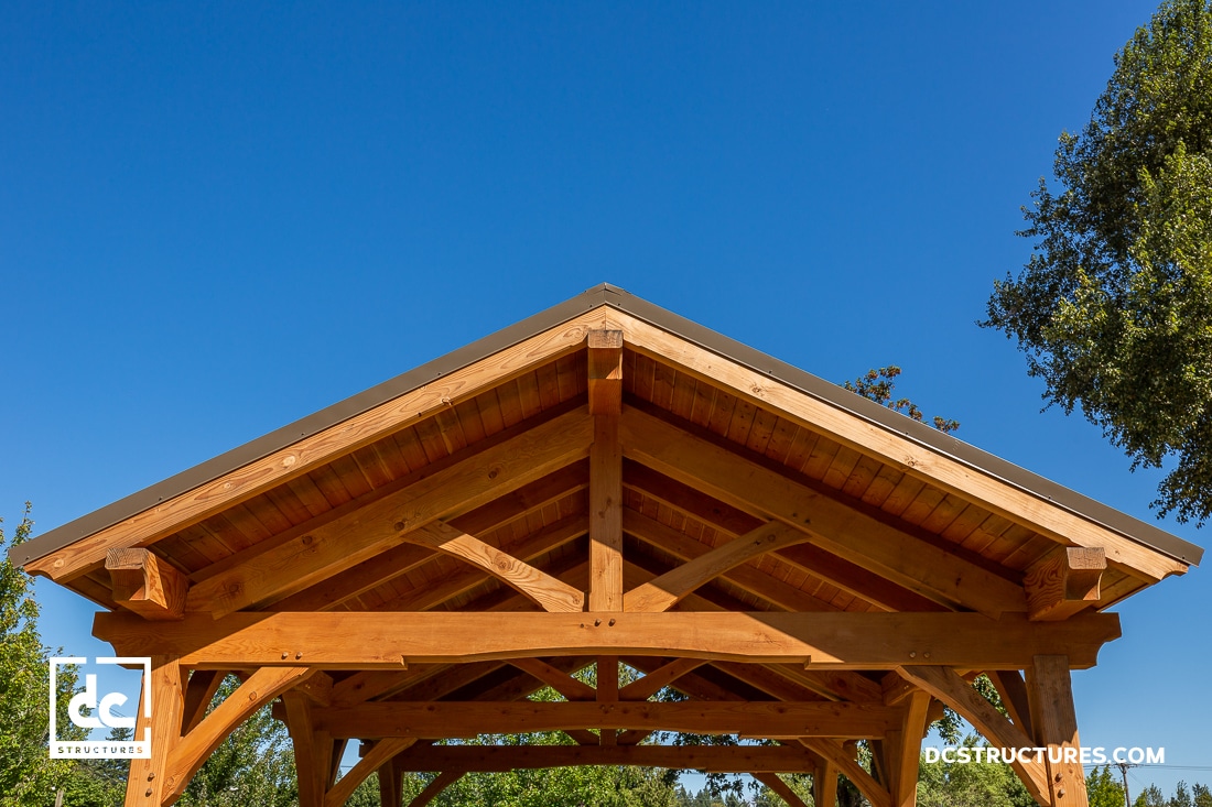 A close-up view of a wooden pavilion roof structure with exposed beams against a clear blue sky, surrounded by greenery.