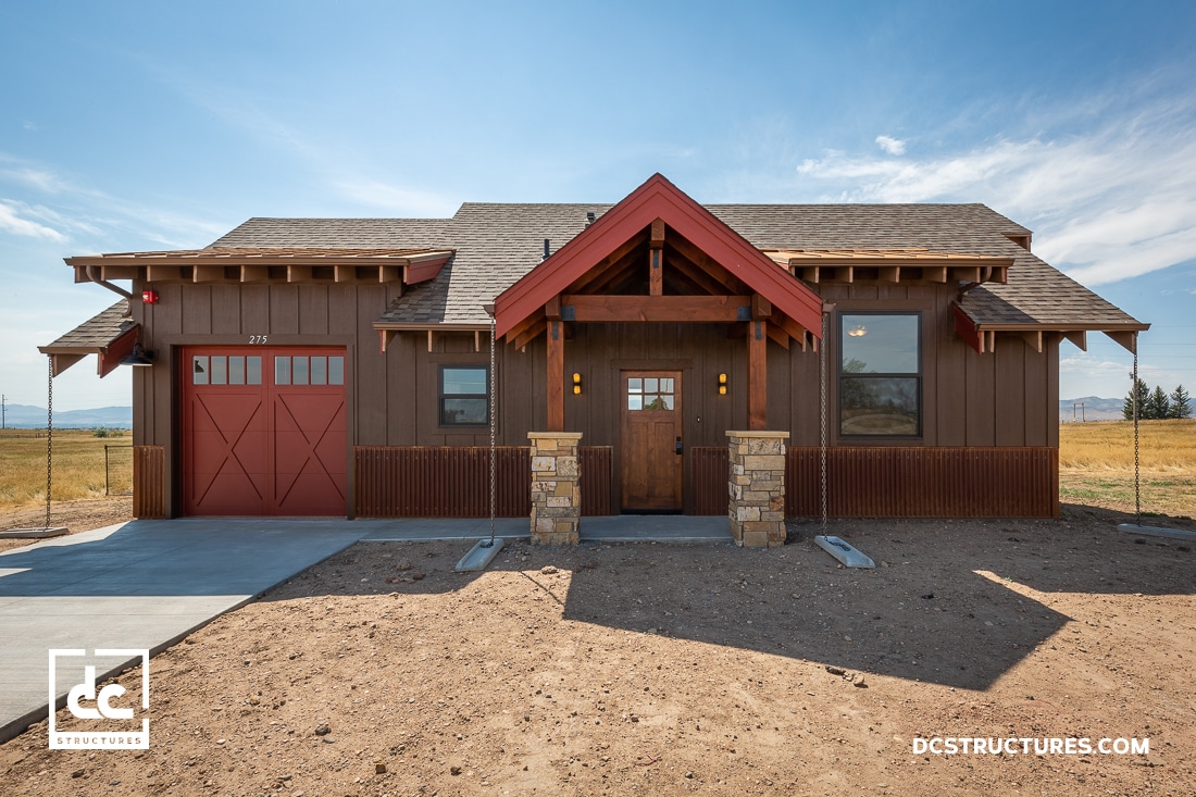 A brown and red wooden house with a large garage door and triangular roof entrance. The house has stone pillars supporting the porch area. It's set against a backdrop of open fields and a partly cloudy sky.