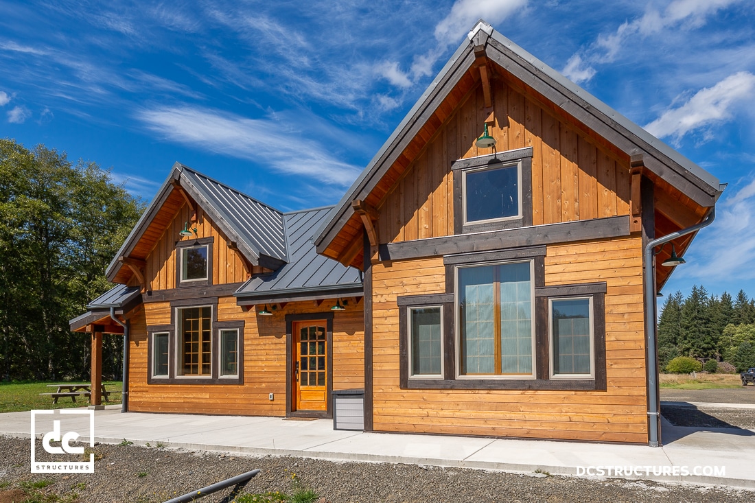 A rustic wooden house, reminiscent of modern cabin kits, features a metal roof, large windows, and a spacious porch. Bathed in sunlight beneath clear skies and surrounded by lush greenery, the scene is complete with a logo in the bottom left corner and text in the bottom right corner.