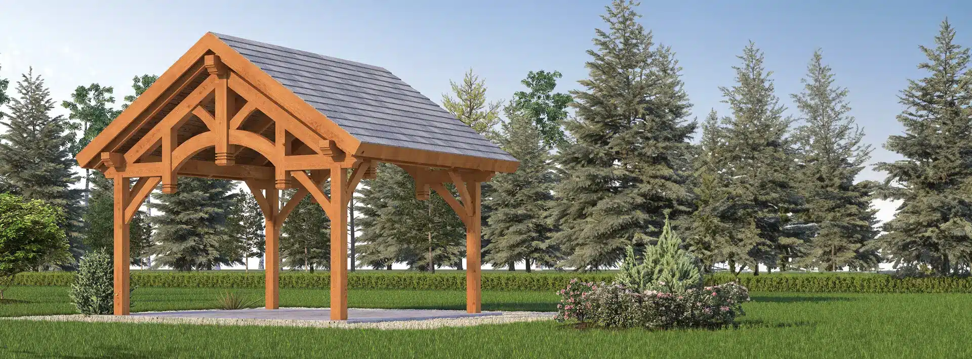 A wooden pavilion with a gabled roof sits on concrete in a grassy area, surrounded by bushes and tall evergreens under blue sky.