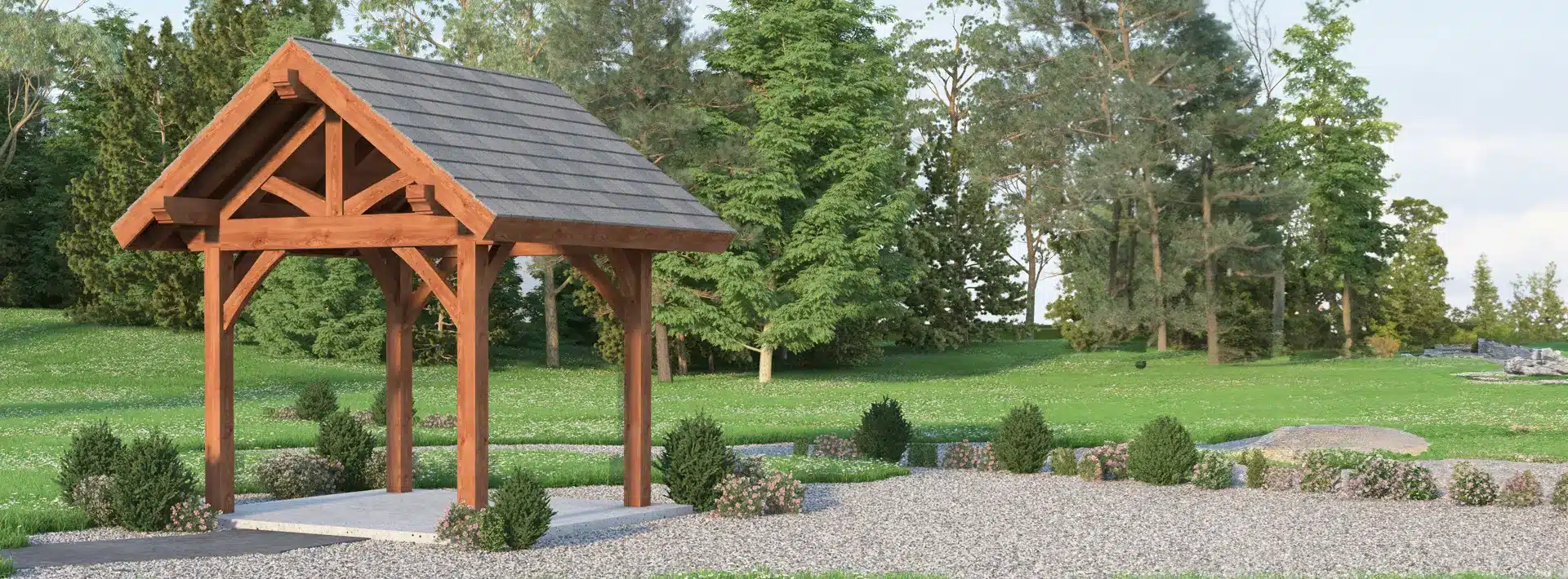 A wooden gazebo with a shingled roof stands on gravel, surrounded by bushes, green grass, and tall trees under a clear sky.