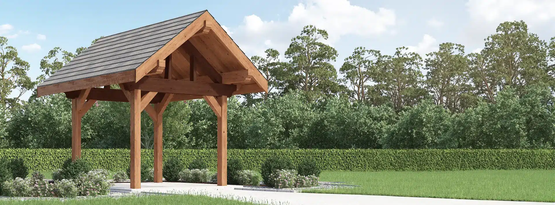 A wooden pavilion with a pitched roof stands on a concrete path, surrounded by green hedges, bushes, grass, trees, and blue sky.