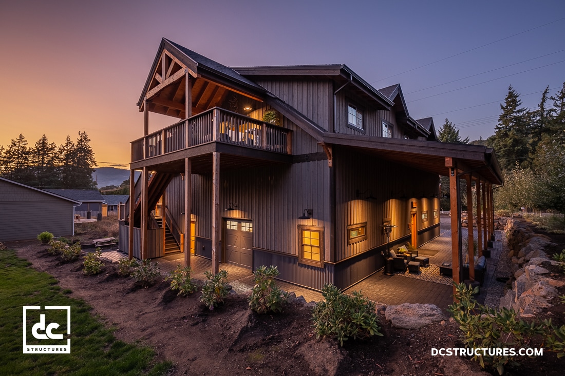 A modern two-story barn-style house with large windows and wooden beams, lit warmly at dusk. Surrounded by a landscaped garden and trees, the house has a wraparound porch. A logo "DC Structures" is visible in the foreground.